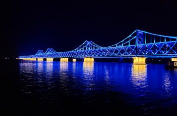 A picture of the rail bridge conencting Dandong and Sinuiju at night. The blue and yellow lights on the bridge reflect off the Yalu River.