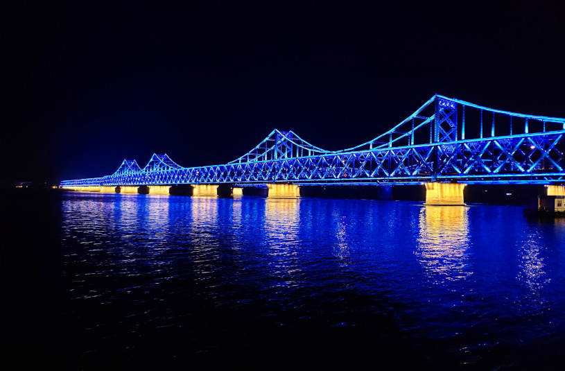 A picture of the rail bridge conencting Dandong and Sinuiju at night. The blue and yellow lights on the bridge reflect off the Yalu River. 