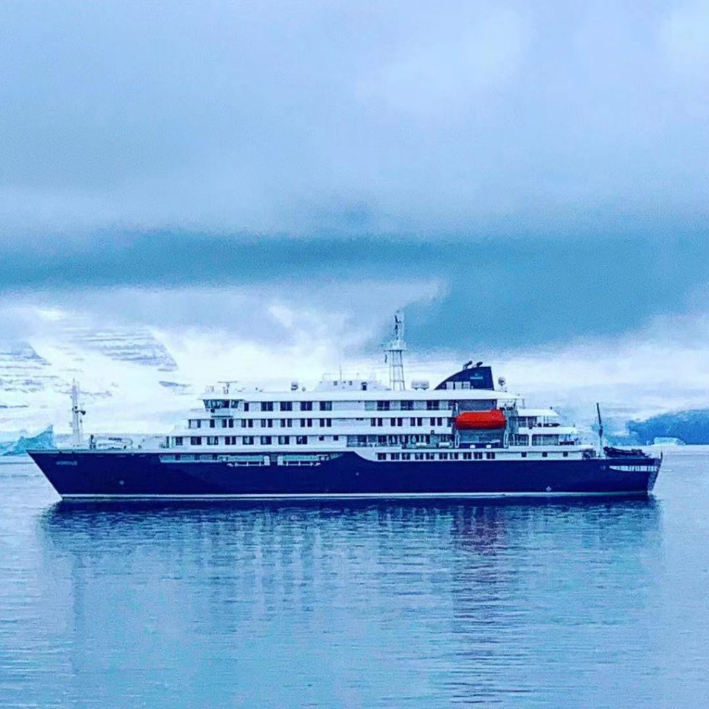 The ship that took us on our Greenland cruise sits in the still ocean under a cloudy sky.