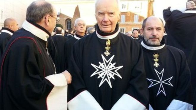 Three men dressed in the black robes of the Sovereign Military Order of Malta stand in the street.