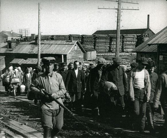 A black and white image of Soviet deportees being escorted by armed guards. 